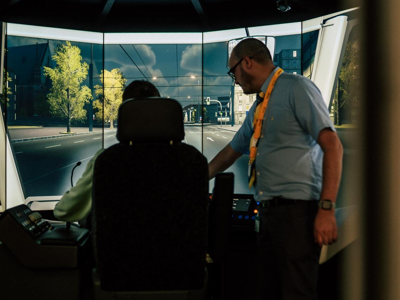 Eine Person sitzt in einem Fahrersitz - zu sehen von hinten. Vor der Person sind Bildschirme in Form einer Straßenbahnscheibe, ein Fahrlehrer erläutert die Armaturen 