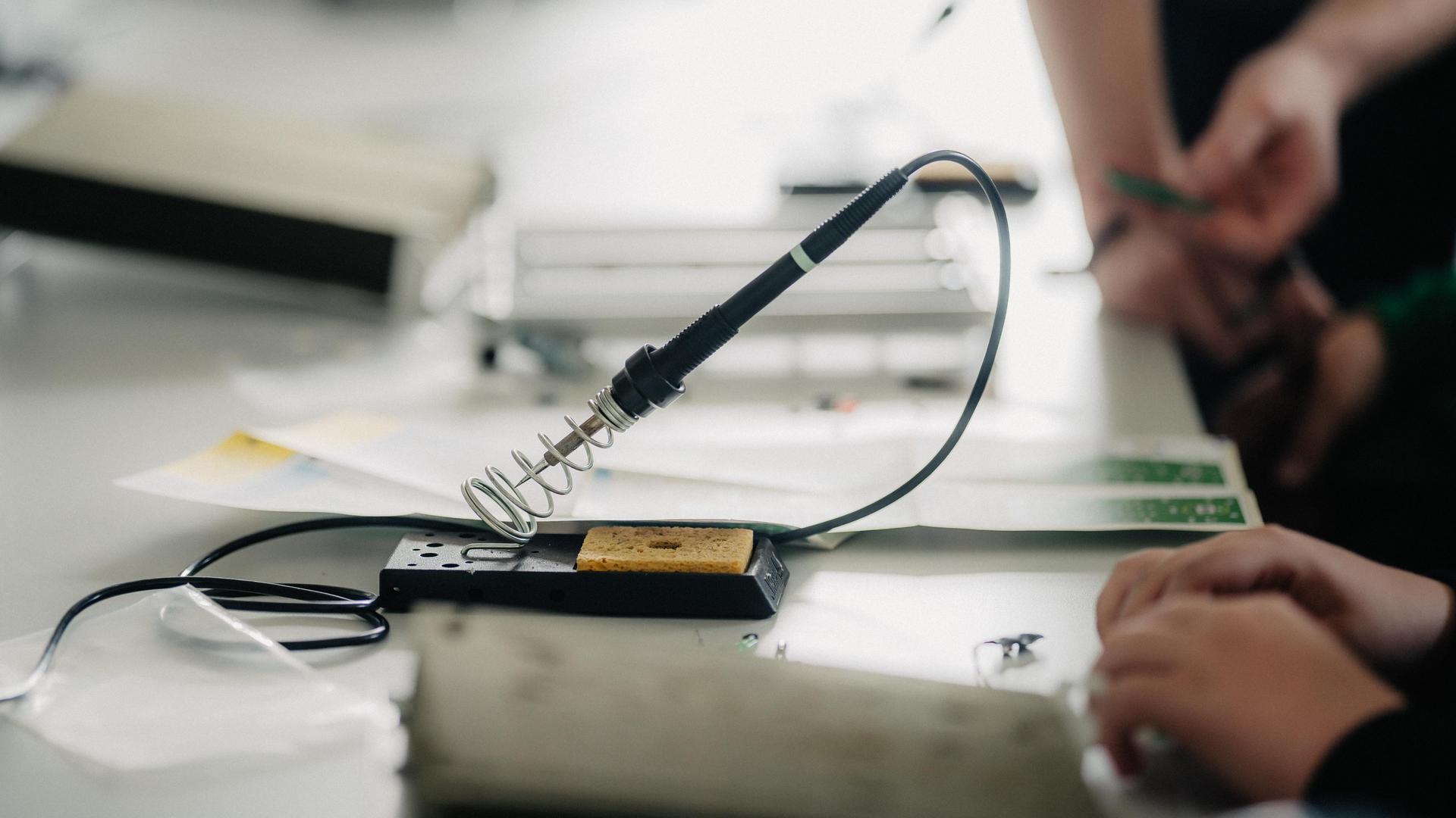 Ein Lötkolben steht in einem speziellen Ständer auf einem Werktisch, im Vordergrund sind Hände zu sehen