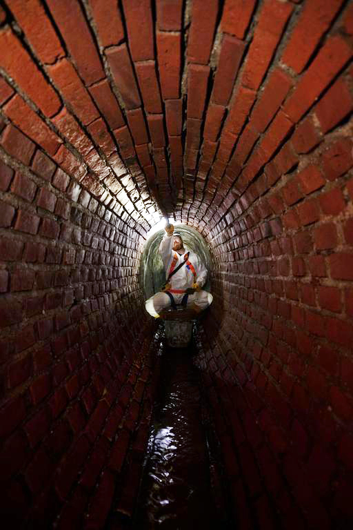 In einem lang, dunklen gemauerten Abwasserkanal sitzt in einiger Entfernung ein Bauarbeiter in weißer Schutzkleidung und arbeiten an den Kanalwänden. Er muss sitzen, weil der Kanal zum Stehen zu niedrig ist.