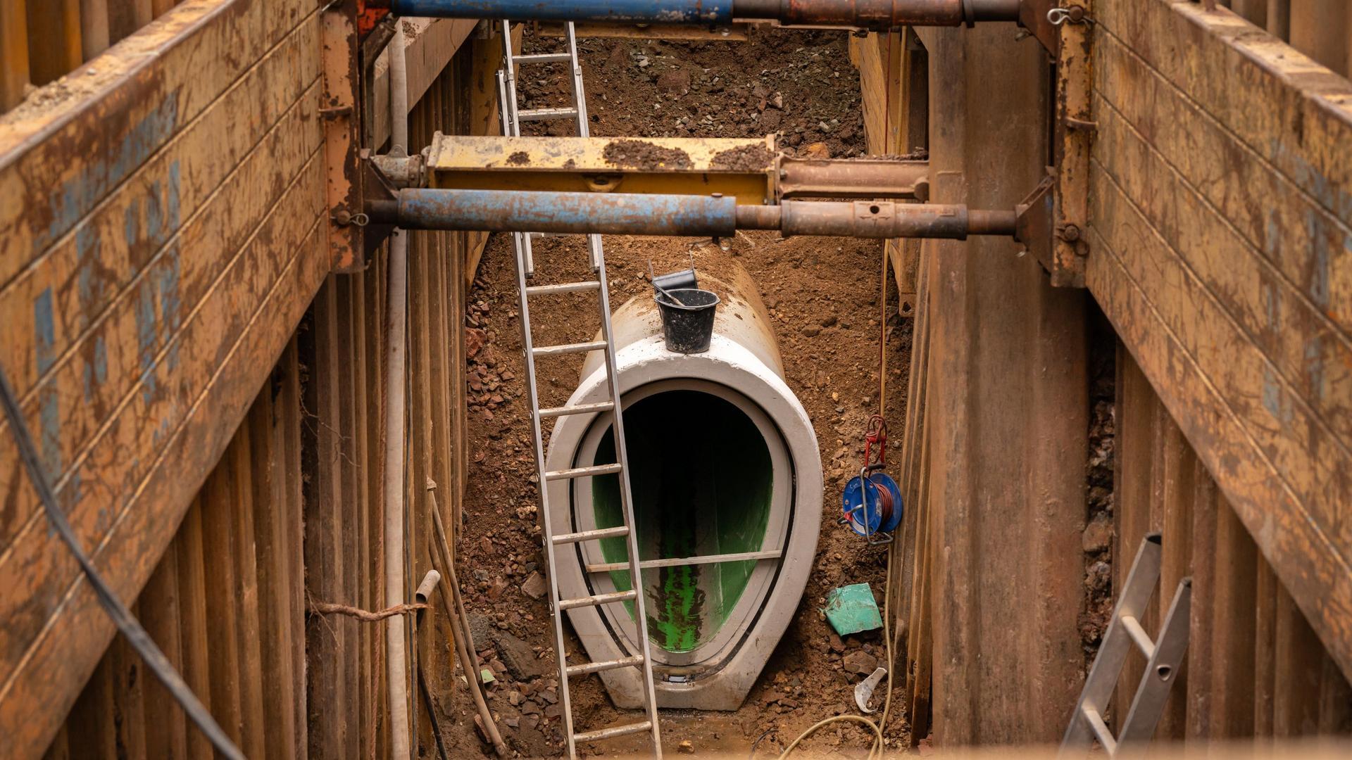 In einer tiefen Baugrube, die durch Spundwände gesichert wird, ist ein eiförmiges Kanalstück zu sehen. Es ist zu erkennen, dass dieses Kanalstück neu eingebaut wurde. 